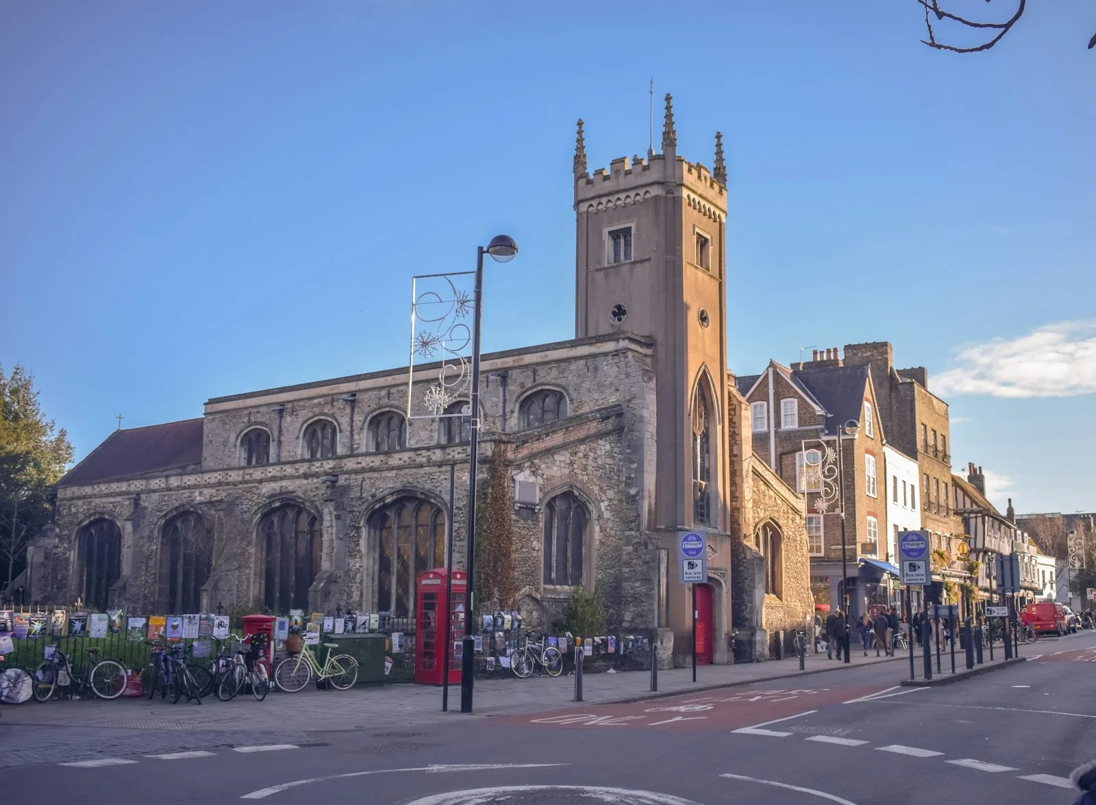A Cambridge street with a stone church, bicycles, a red phone box and shops